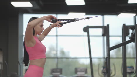 A-brunette-woman-in-a-pink-suit-pulls-a-rope-in-a-crossover-trainer-to-her-chest.-Back-workout-in-a-trainer.-Professional-woman-instructor